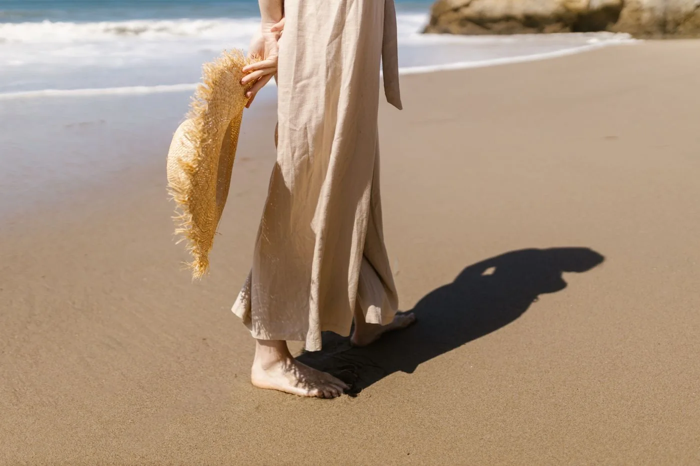 Woman wearing light trousers and holding a straw hat on a sandy beach.